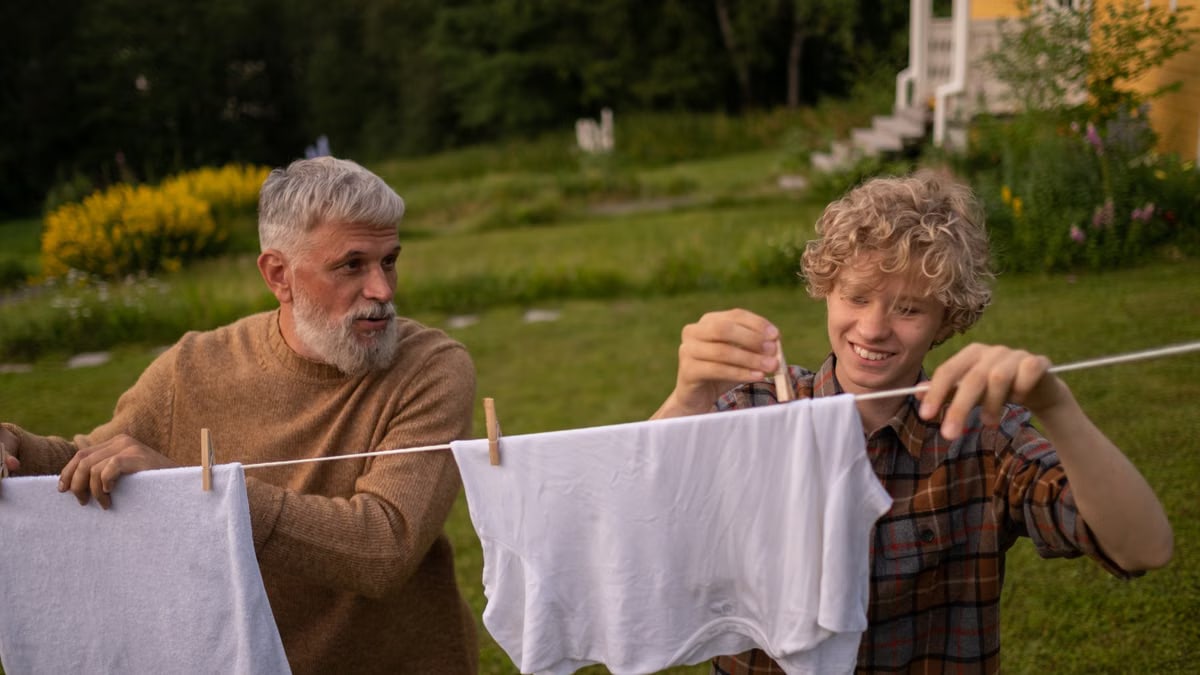 Elderly man and teenage boy joyfully hanging laundry in a sunny garden setting.